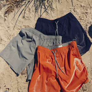Three sandy swimshorts - in navy blue, light grey and bright orange - laid out on beach sand with dune-grass shadows.