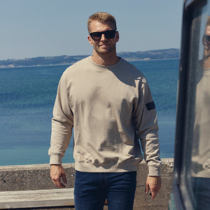Man wearing a light beige sweatshirt and dark blue jeans, standing by the waterfront with a scenic ocean view and his reflection visible in a neaby car window.
