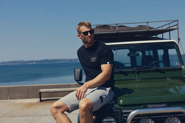 Man wearing a dark blue Compass Classic t-shirt with a small text print on left chest and sunglasses sitting on a dark green vintage Landrover Defender near the water, with a coastal background and blue sky.